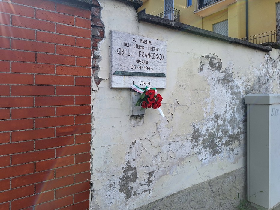 Lapide commemorativa a Gibelli, decorata con fiori, su muro di una struttura rustica.