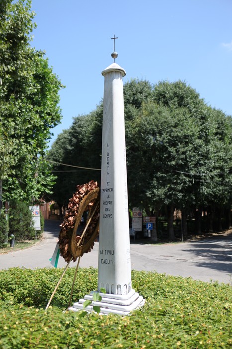 Monumento ai caduti circondato da vegetazione, con una corona di fiori e il cielo blu sullo sfondo.