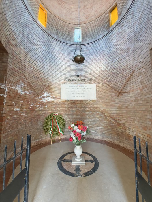 Interno del Sacrario ai Caduti Partigiani di Piumazzo, con decorazioni floreali e una lapide commemorativa.