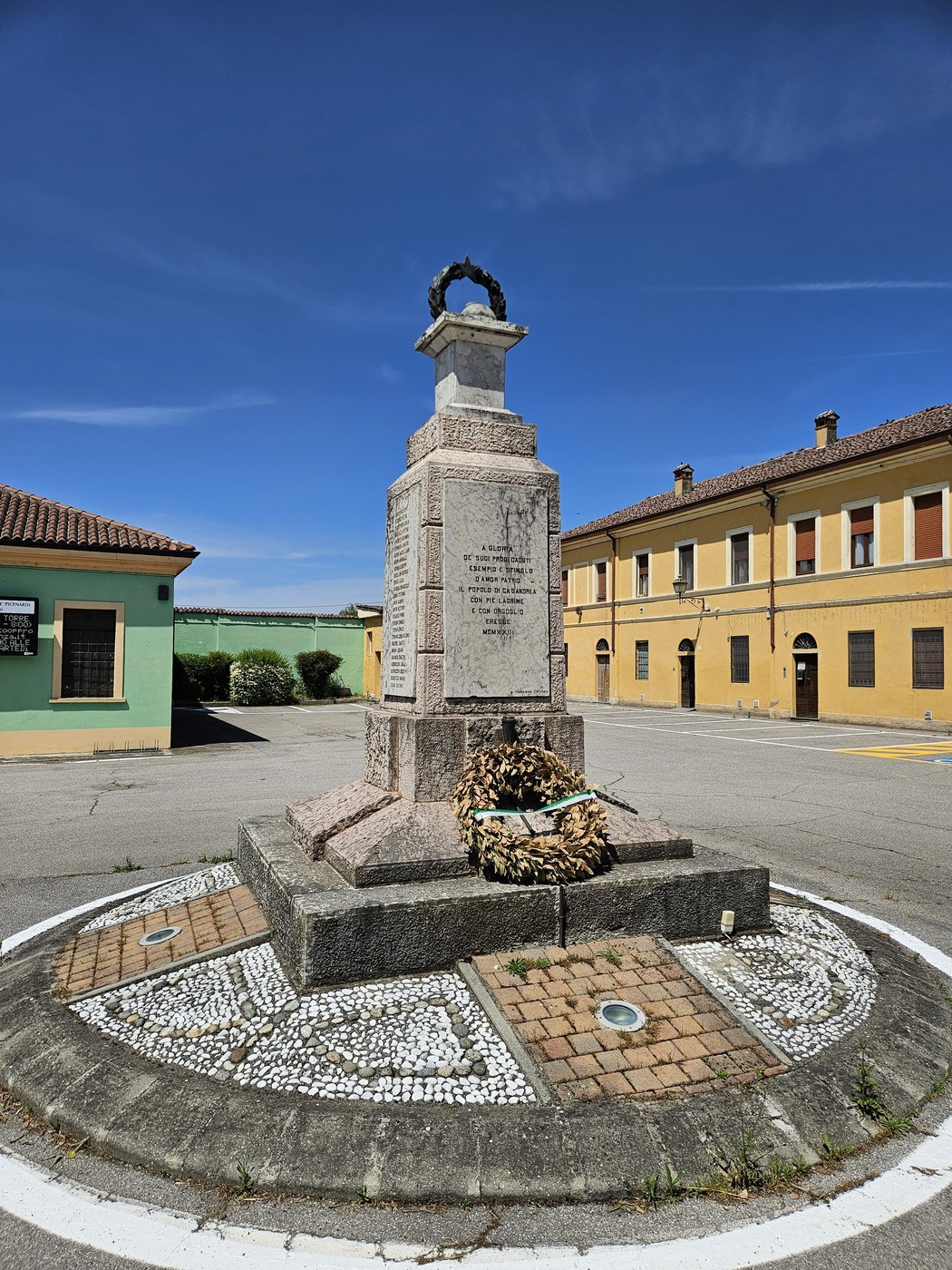 Monumento ai Caduti di Cà d'Andrea, circondato da un'area curata e edifici colorati sullo sfondo.