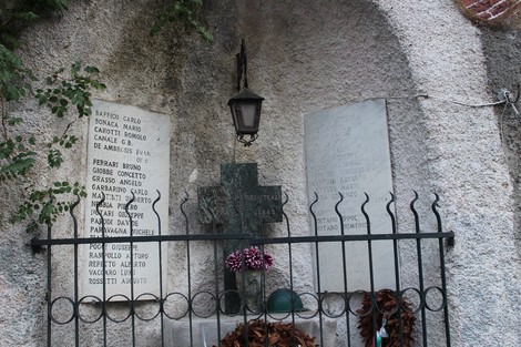 Edicola commemorativa con nomi di caduti, croce metallica e lanterna in un contesto di pietra.