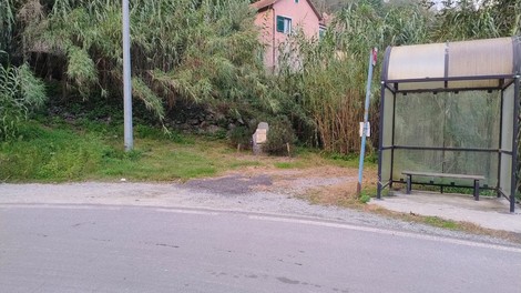 Cippo in un'area verde vicino a una fermata dell'autobus, circondato da vegetazione e edifici.