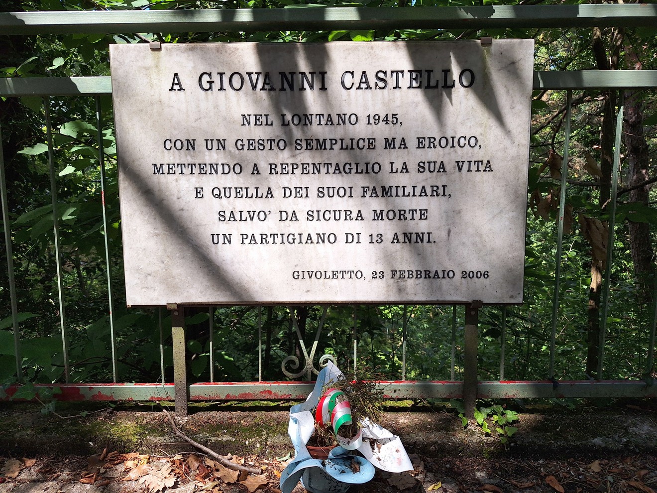 Lapide commemorativa dedicata a Giovanni Castello, con un omaggio floreale in un contesto naturale.