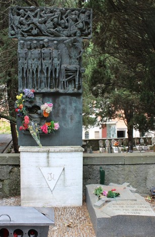 Monumento ai deportati, con bassorilievi e fiori commemorativi, in un contesto di cimitero.