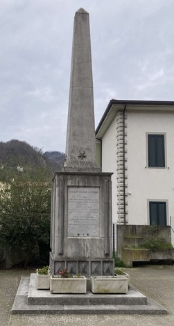 Monumento ai caduti di Vado, obelisco di pietra con dedicato e fiori, circondato da edifici e natura.