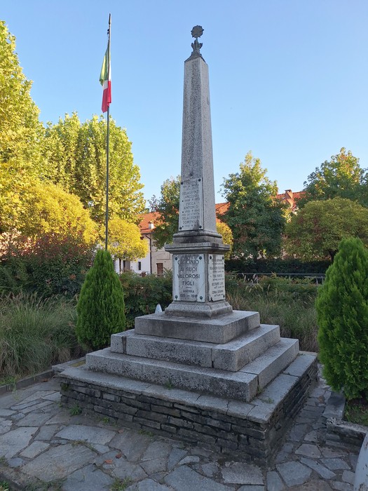 Obelisco in pietra su basamento a gradoni con iscrizione, in una piazza con alberi e bandiera italiana.