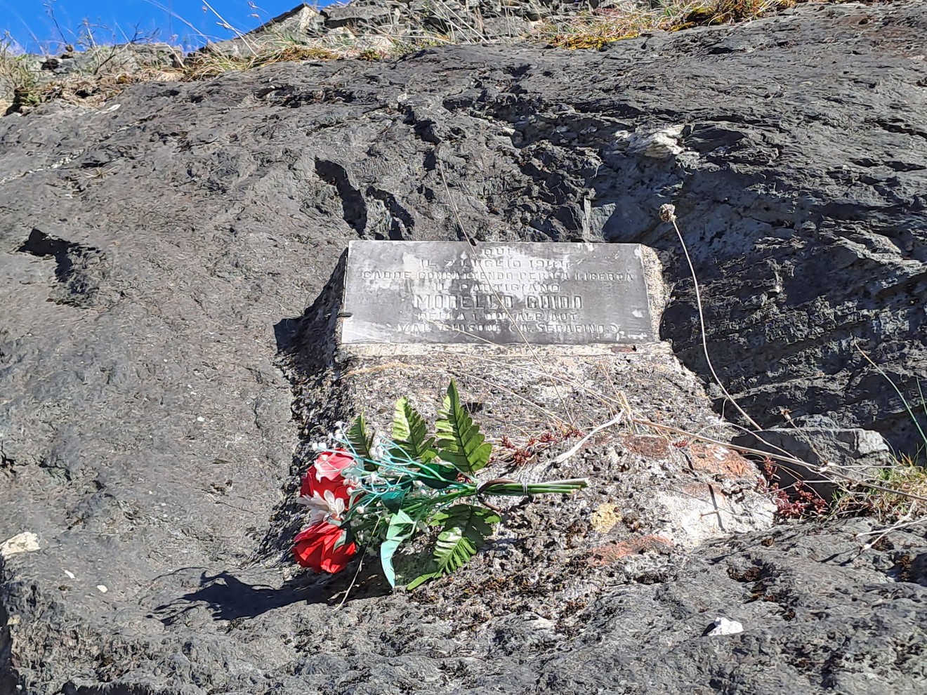 Lapide commemorativa dedicata a Morello, posizionata su un terreno roccioso con fiori decorativi.