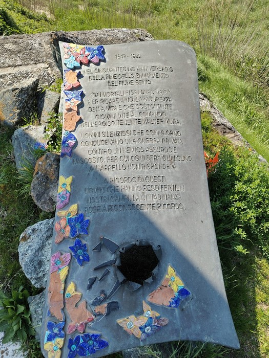 Lastra commemorativa agli sminatori, decorata con fiori e insetti colorati, posta su un terreno naturale.