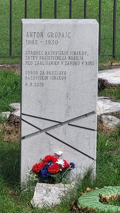 Lapide in pietra chiara con iscrizione e croce incisa, fiori deposti alla base, su prato erboso.