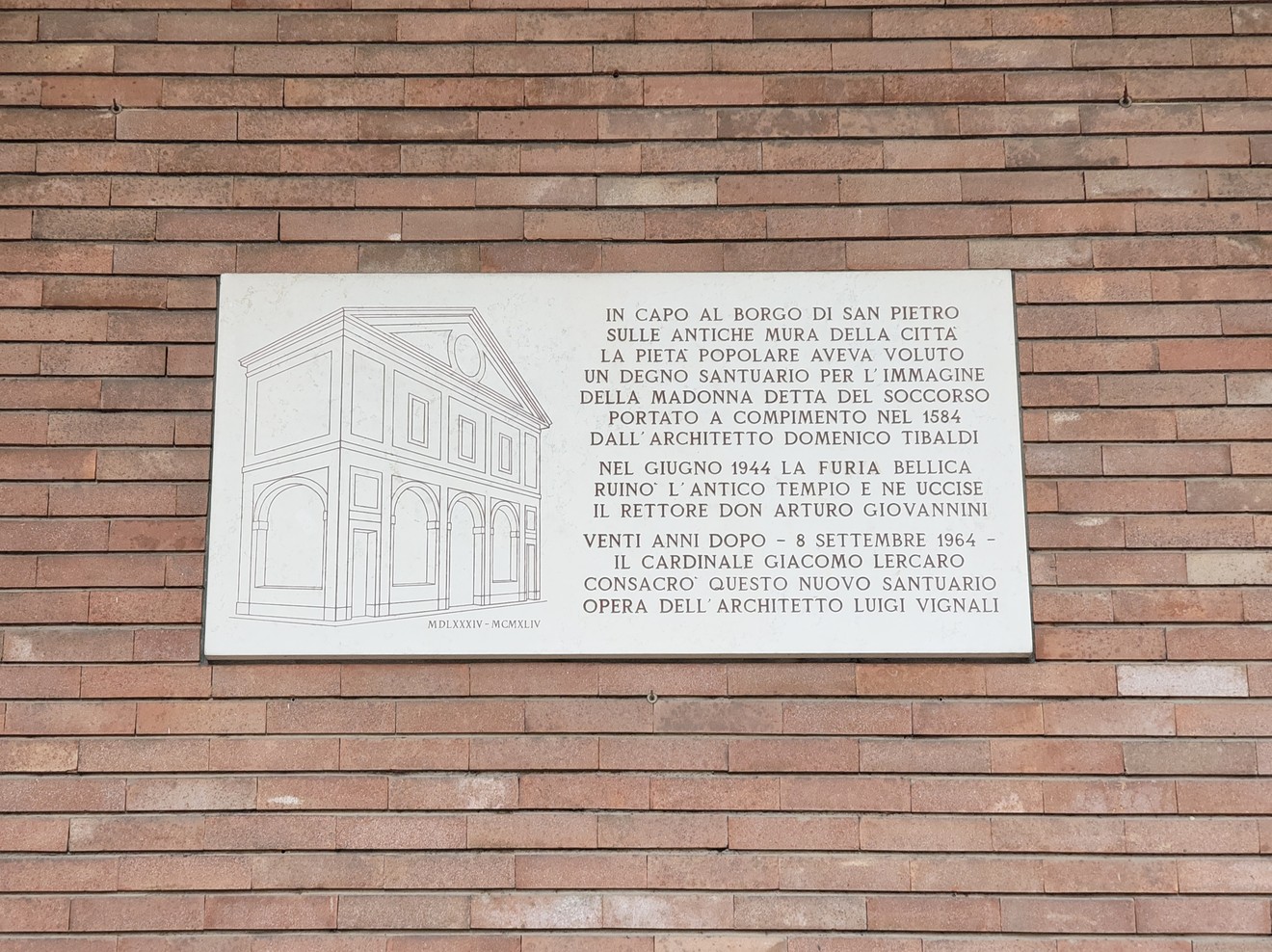 Lapide bianca con bassorilievo della chiesa e lunga iscrizione, su parete di mattoni a vista.