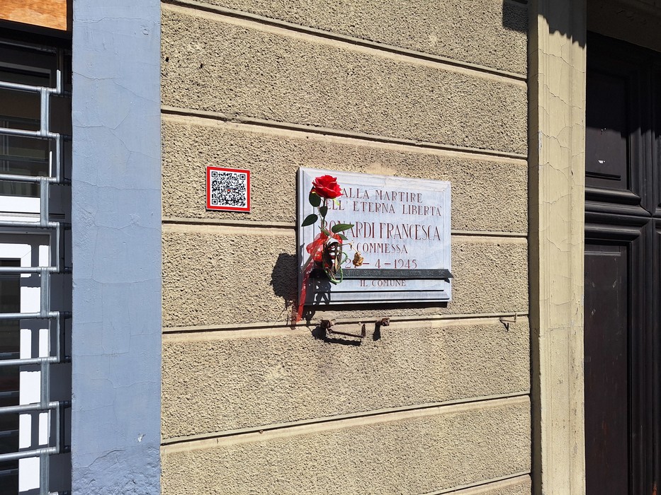 Lapide in marmo su parete esterna, con iscrizione e un fiore rosso appoggiato sotto il testo.