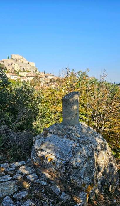 Cippo in pietra con targa incisa su masso, tra la vegetazione, con borgo collinare sullo sfondo.