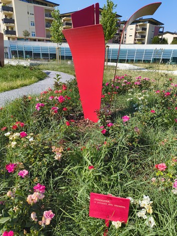 Installazione artistica in un giardino fiorito, con opere rosse e un'atmosfera primaverile.