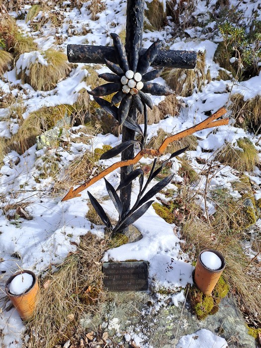 Croce in ferro battuto con stella alpina, targa incisa e due portalumini, tra erba e neve.