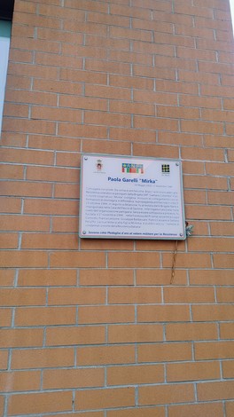 Targa commemorativa su un muro di mattoni, dedicata a Paola Garelli, con informazioni storiche.