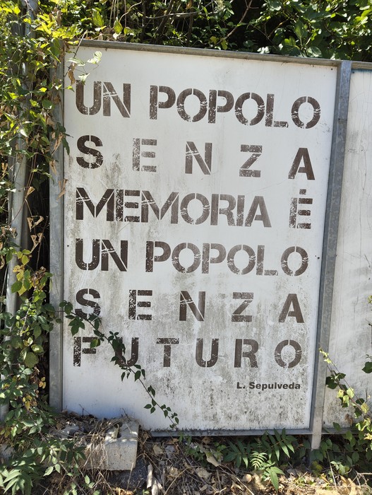 Cartello metallico con iscrizione, montato su pali tra la vegetazione lungo un sentiero.