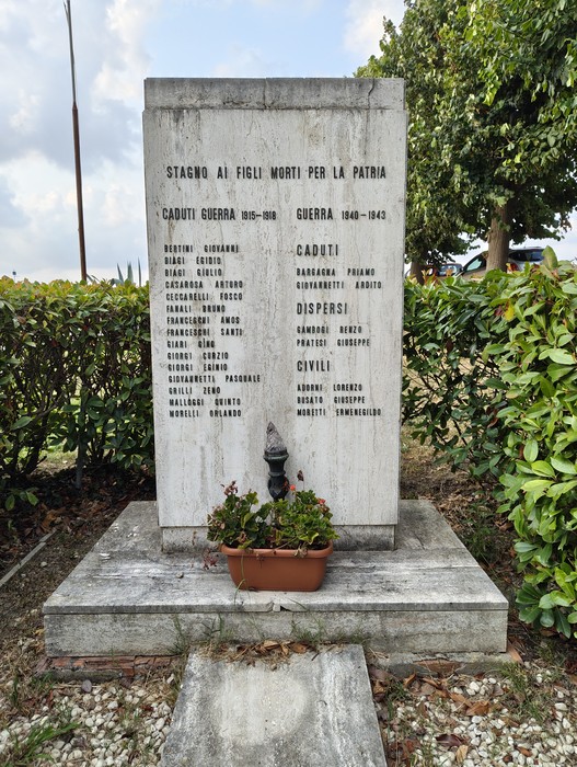 Stele in pietra con iscrizione e elenco di nomi, circondata da siepi, con vaso di fiori alla base.