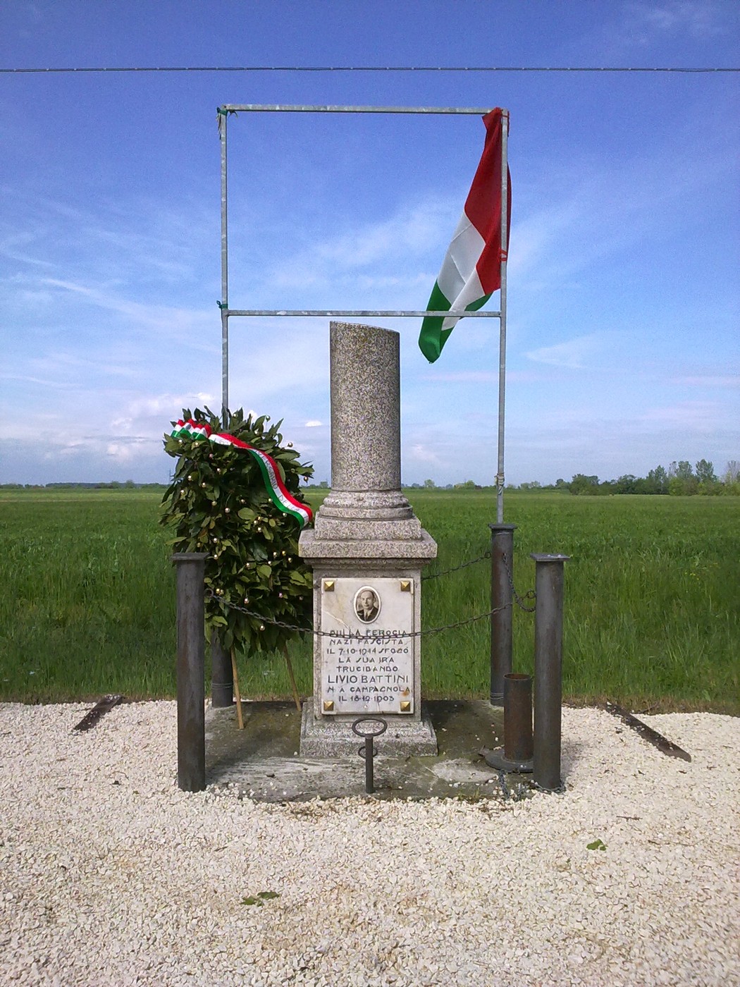 Cippo commemorativo con bandiera italiana e corona d'alloro, immerso nel verde dei campi circostanti.