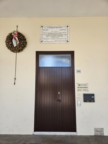 Lapide commemorativa a Fraccon affissa sopra una porta, con una ghirlanda decorativa accanto.