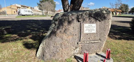 Monumento a Cocco, grande roccia con iscrizione e fiori, circondato da un'area verde e edifici sullo sfondo.