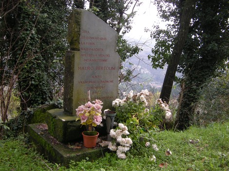 Cippo commemorativo dedicato a Mauro Bertolini, circondato da fiori e vegetazione, con vista panoramica.