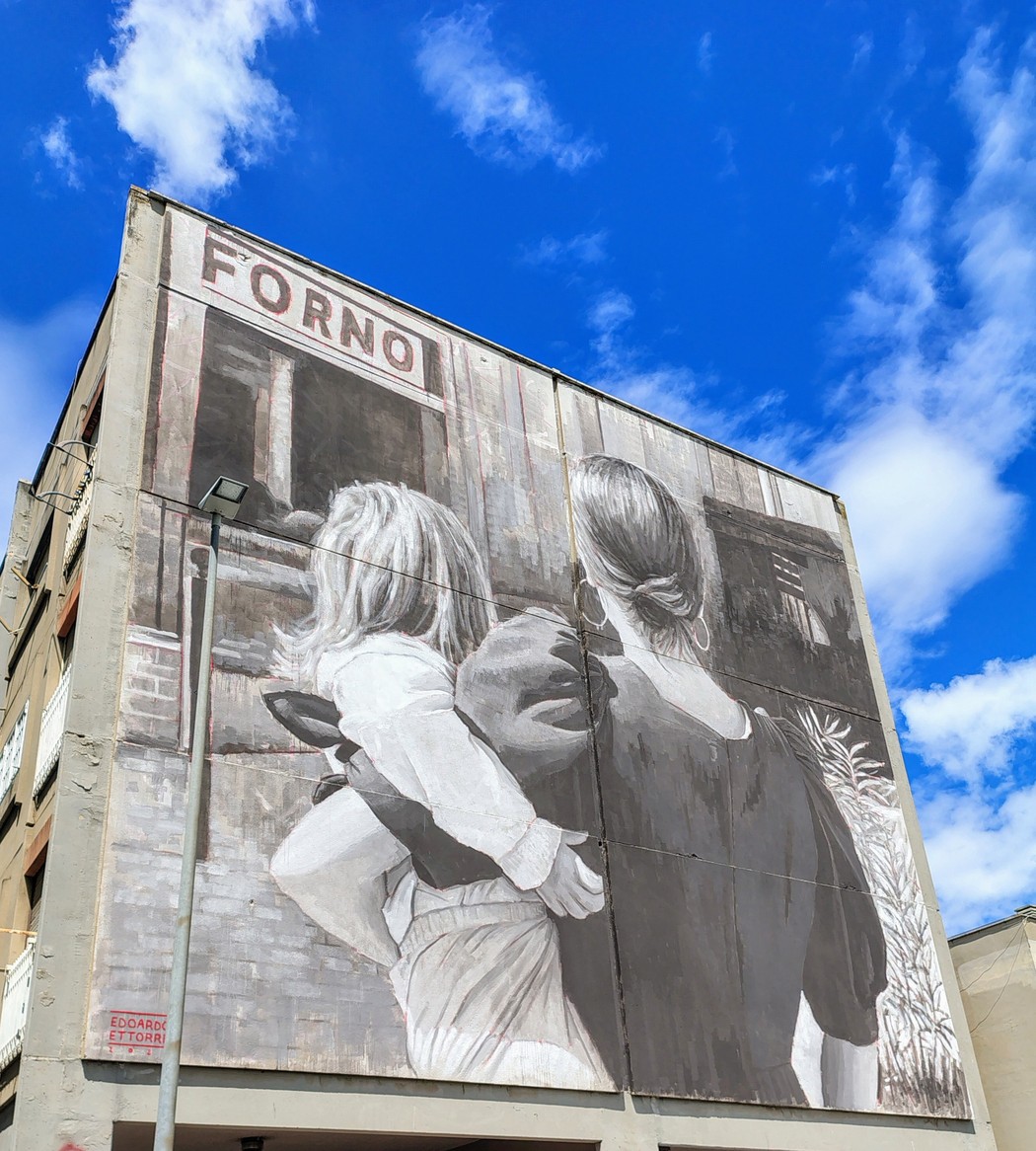 Murale che ritrae due figure femminili su un edificio, con cielo azzurro e nuvole bianche sullo sfondo.