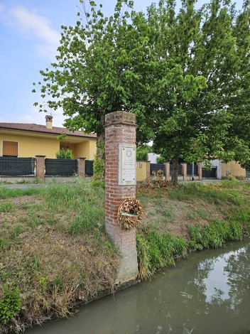 Cippo commemorativo a Cavagnari, immerso nel verde, affiancato da una vegetazione rigogliosa e un canale adiacente.