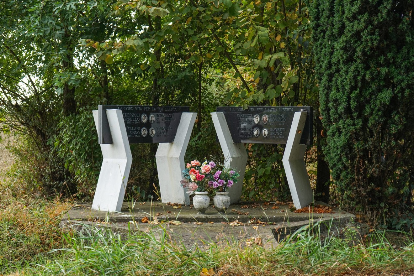 Monumenti commemorativi circondati da vegetazione, con fiori e ritratti in memoria di Pederzoli e altri.
