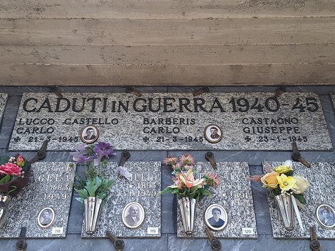 Lapide in marmo con iscrizione e fototessere dei caduti, sotto fiori in vasetti metallici alla parete.