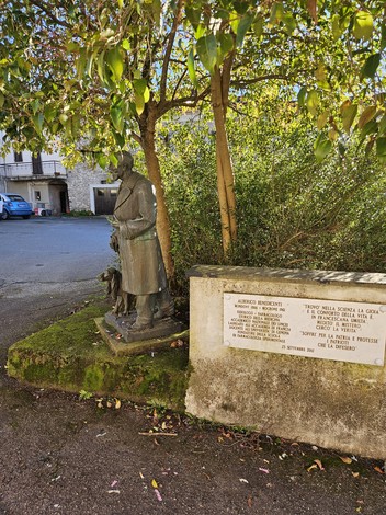 Monumento a Benedicenti, con statua e base informativa, immerso nella vegetazione.