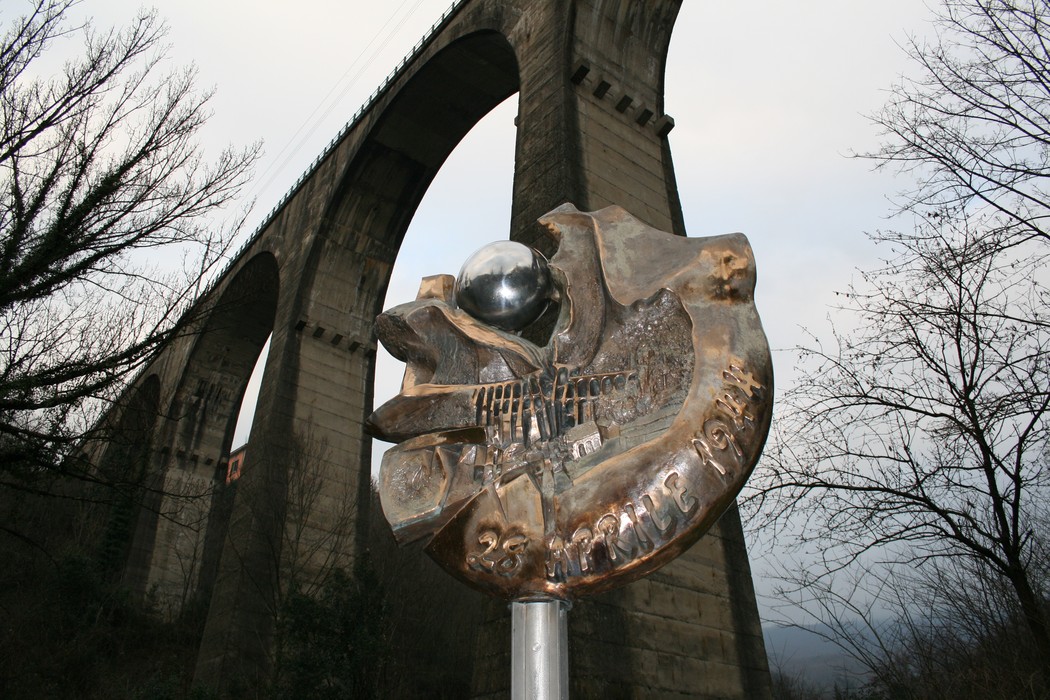 Monumento commemorativo ai bombardamenti della Porrettana, con una scultura e un viadotto sullo sfondo.