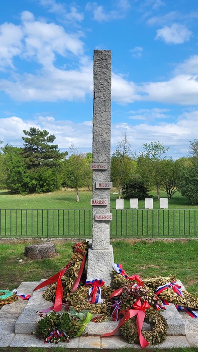 Cippo in pietra con targhe incise, circondato da corone di fiori, in un parco recintato.