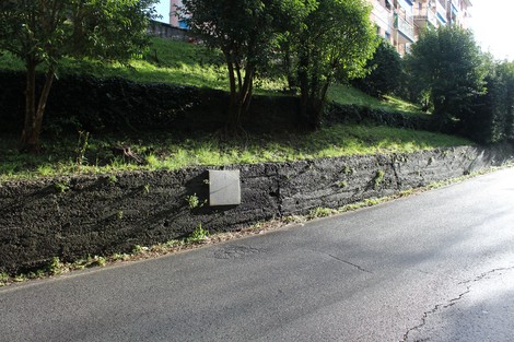 Lapide affissa a un muro di pietra, circondata da vegetazione e visibile lungo una strada.