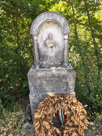Cippo in pietra con bassorilievo di una figura e iscrizione, corona di alloro ai piedi, nel bosco.
