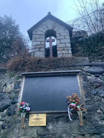 Lapide commemorativa di Malatrait con croce sopra, adornata da fiori, immersa in un ambiente naturale.