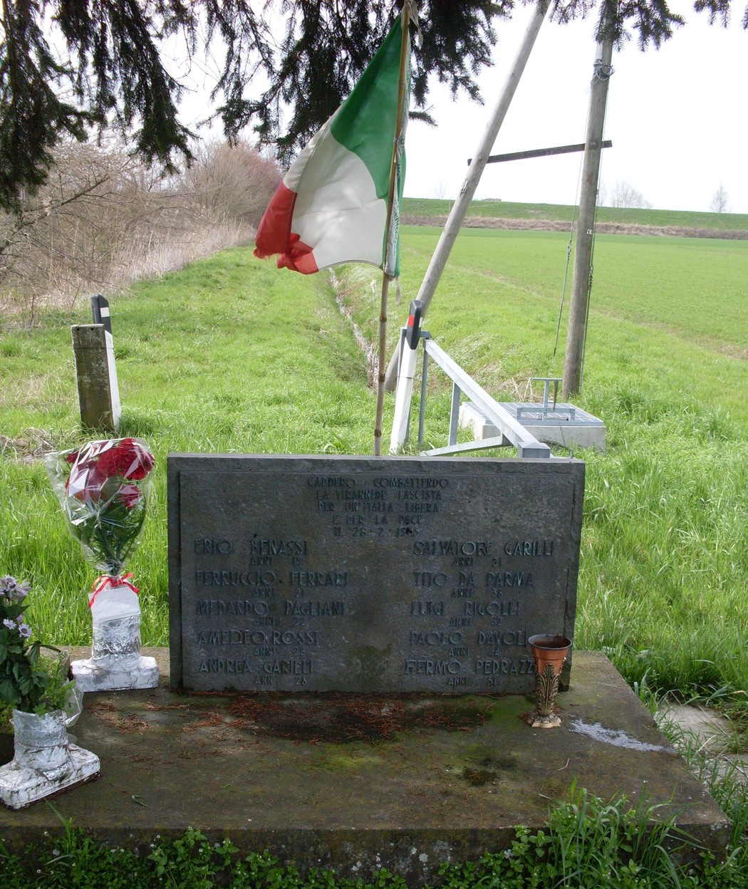 Monumento commemorativo per l'eccidio di Cadelbosco, con fiori e bandiera italiana, in un contesto naturale.
