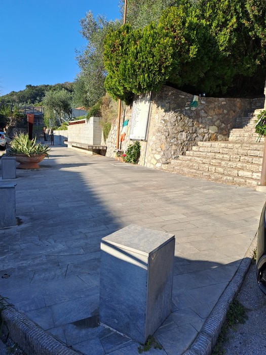 Lapide commemorativa con iscrizione affissa a un muro in pietra, lungo una piazza pavimentata.