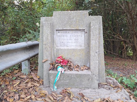 Cippo in pietra con iscrizione e mazzo di fiori ai piedi, lungo una strada tra alberi.