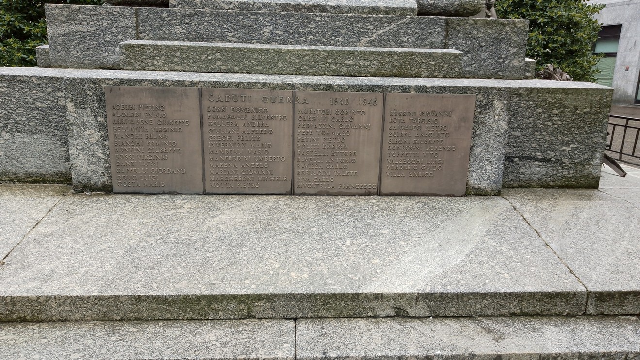 Monumento ai caduti con lastre commemorative incise su pietra, circondato da vegetazione e ambiente urbano.