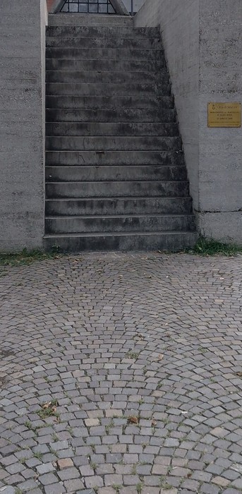 Scalinata di cemento che conduce al "Monumento ai Partigiani", circondata da pavimentazione in ciottoli.