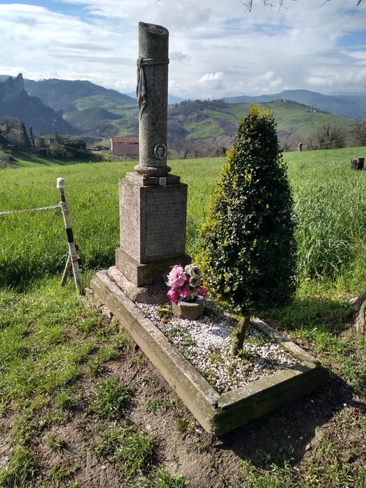Cippo commemorativo in un paesaggio collinare, con fiori e una pianta decorativa in un ambiente verdeggiante.