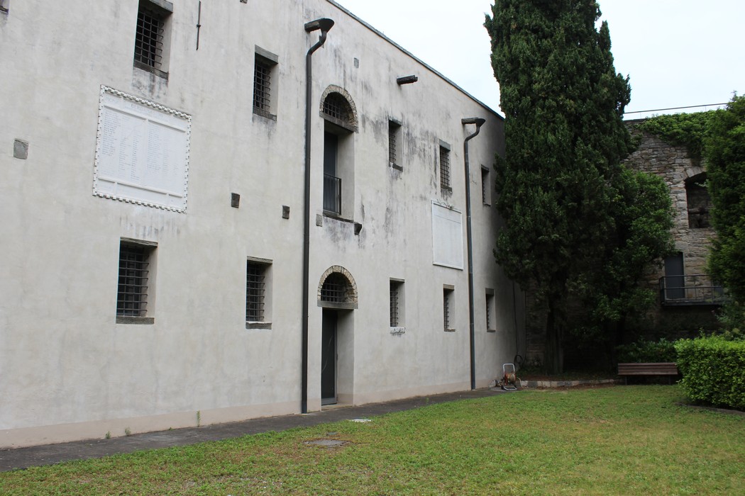 Stele commemorativa in un'area verde, affiancata da edifici storici e alberi.