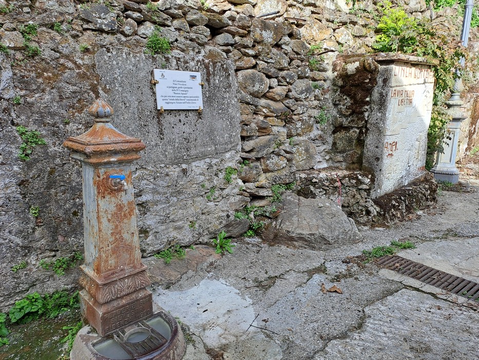 Lapide con iscrizione su parete in pietra, accanto a una vecchia fontanella in ghisa arrugginita.