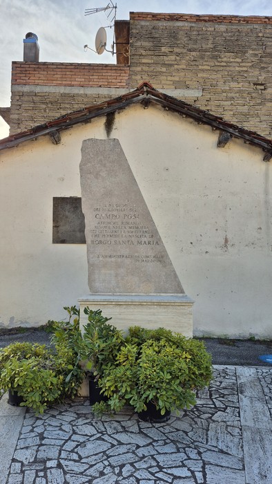 Monumento al Campo PG54, con una struttura in pietra e vegetazione circostante, su sfondo di un edificio.