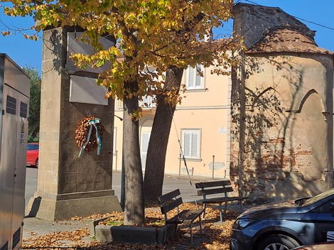Lapide ai caduti di Mezzana decorata con una corona di fiori, circondata da panchine e alberi in autunno.