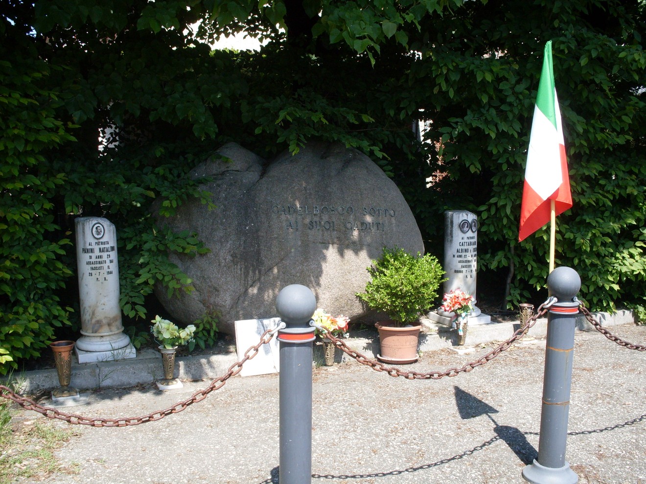 Monumento dedicato ai Cattabiani e a Panini, con fiori e bandiera italiana in un contesto curato e pittoresco.