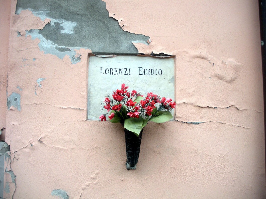Targa commemorativa di Lorenzi con fiori rossi, affissa su una parete danneggiata.