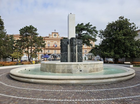 Fontana Monumentale circondata da alberi e una pavimentazione in ciottoli, con uno sfondo architettonico elegante.