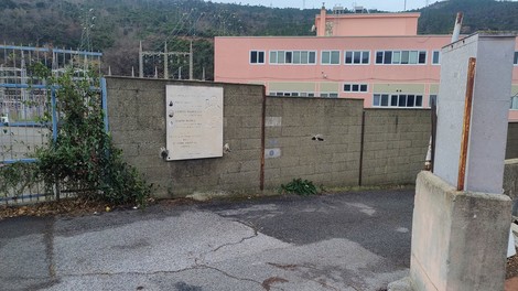 Lapide commemorativa vicino a un muro, con edifici sullo sfondo e vegetazione circostante.