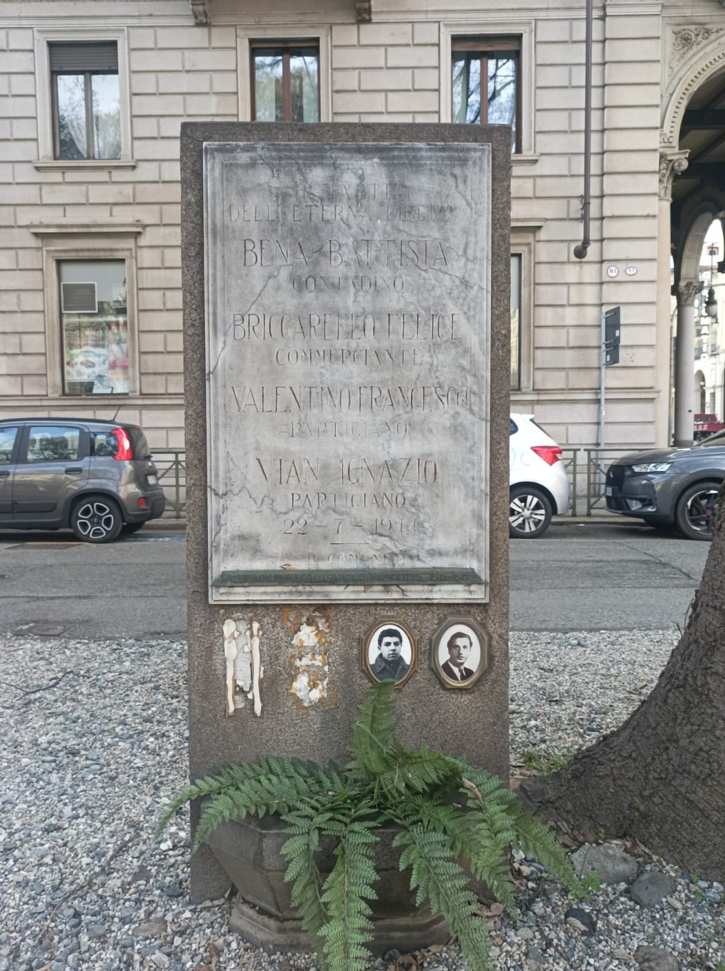 Stele commemorativa con nomi incisi, circondata da elementi naturali e architettura urbana sullo sfondo.